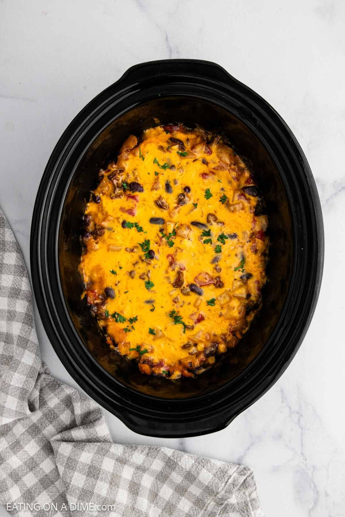 A Crockpot Cowboy Casserole filled with melted cheesy dip, ground meat, and herbs sits on a white marble surface beside a gray checkered cloth.