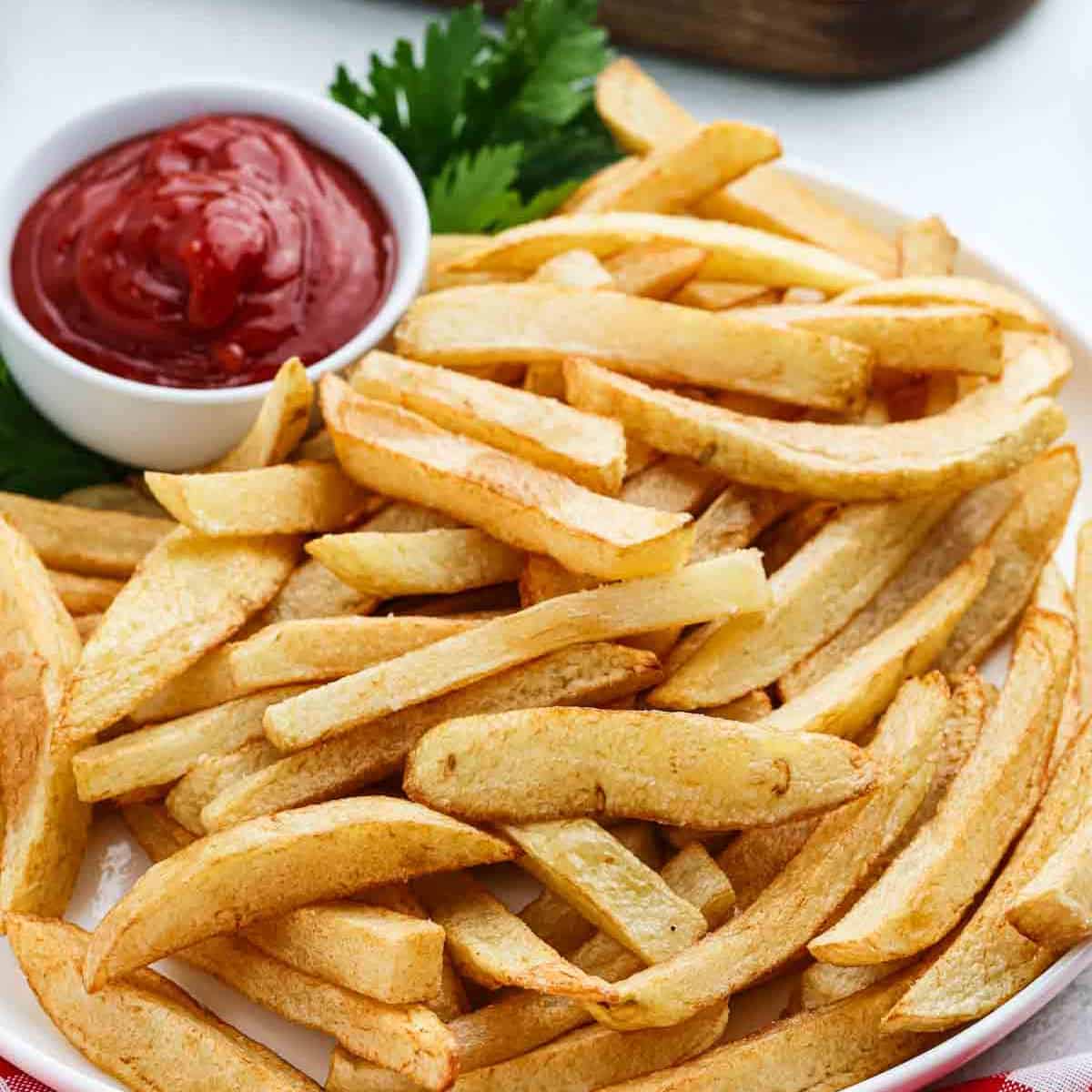 A plate of golden, crispy homemade French fries is served with a small bowl of ketchup and a garnish of fresh parsley in the background.
