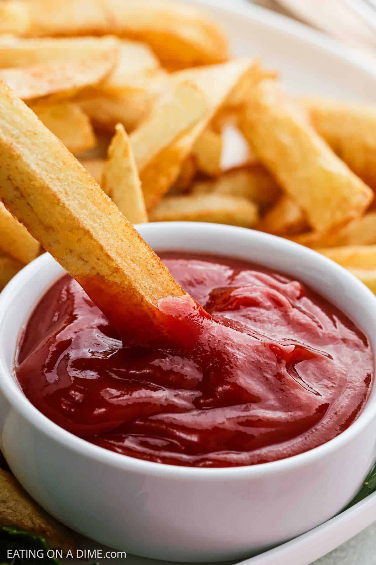 Golden French fry being dipped into a bowl of ketchup, with more french fries in the background on a white plate.