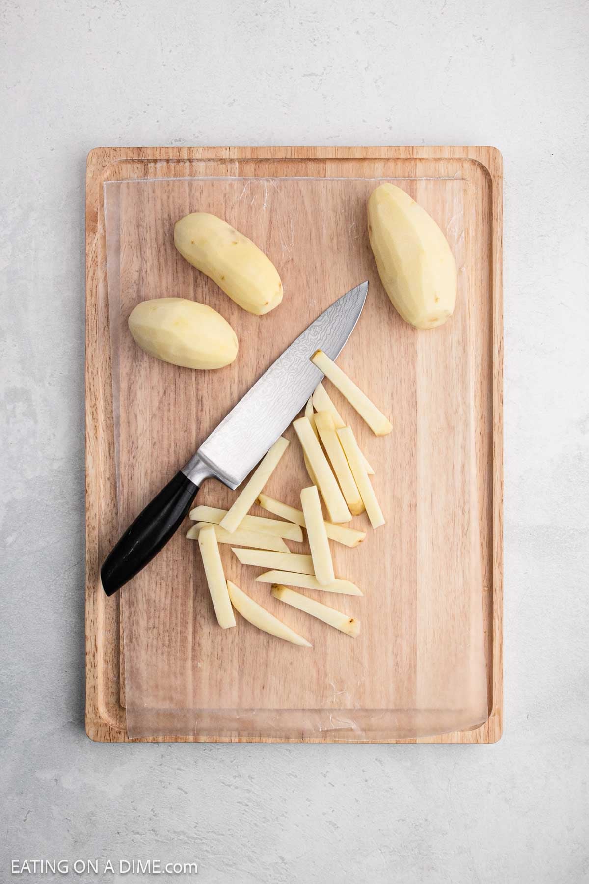 Peeled potatoes on a cutting board with one cut into strips with a large knife on the side.