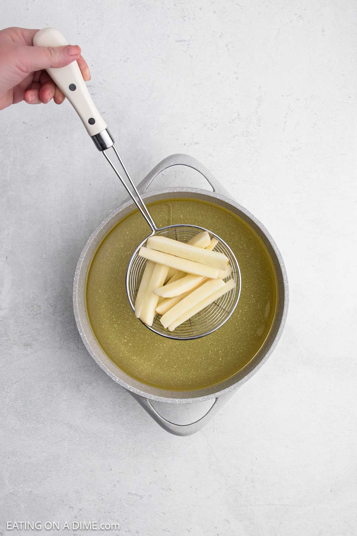 A hand holds a slotted spoon with raw potato sticks above a pot filled with oil.