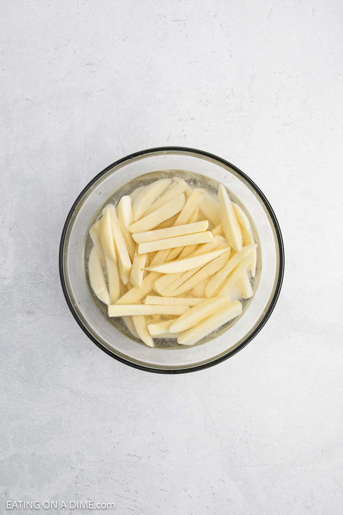A glass bowl filled with raw potato slices soaking in water on a light-colored surface.