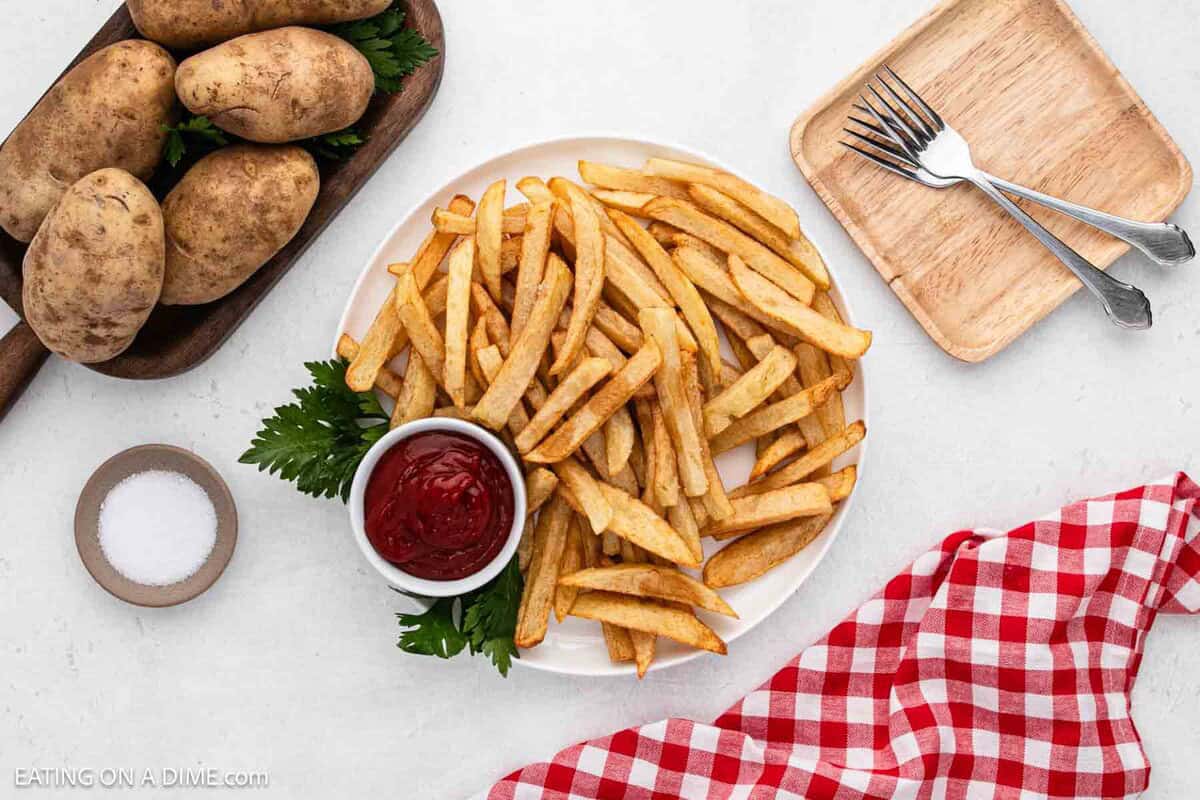 Homemade French Fries on a white plate with a small bowl of ketchup on the side. A wooden platter with whole Russet Potatoes are on the side.