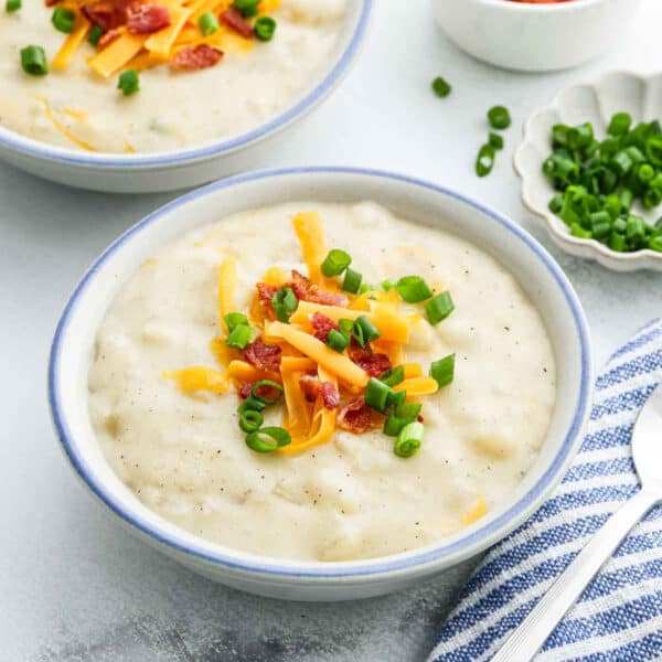 A bowl of creamy Instant Pot loaded baked potato soup topped with shredded cheddar cheese, crispy bacon bits, and chopped green onions sits on a light surface next to a striped napkin and spoon. Another bowl and extra toppings are nearby.
