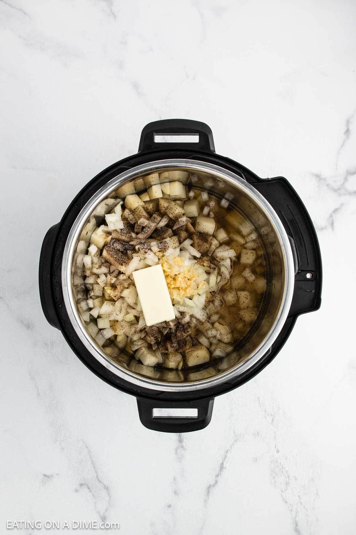 An overhead view of an Instant Pot ready to make loaded baked potato soup, filled with chopped potatoes, onions, minced garlic, seasonings, and a block of butter. The pot sits on a white marble surface.
