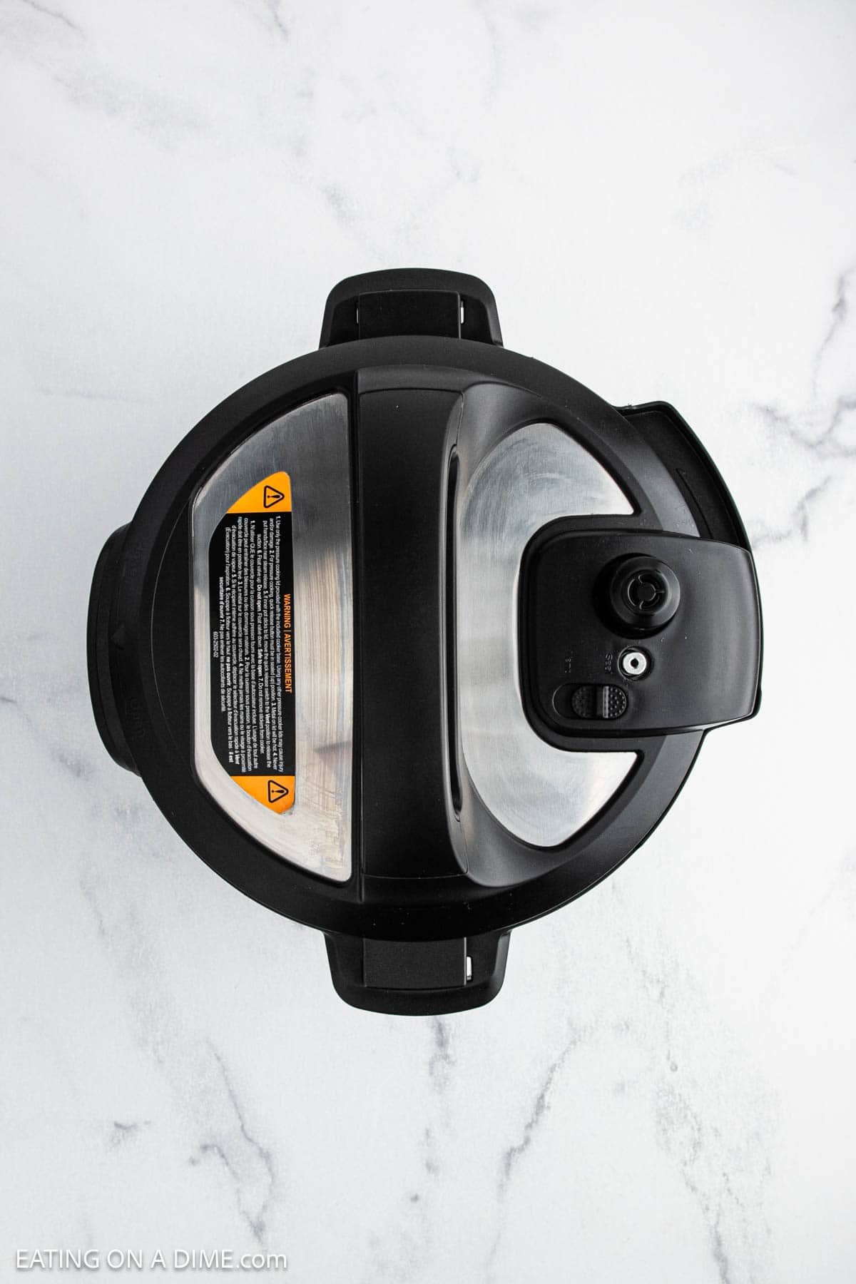 Top-down view of a black and silver pressure cooker with a closed lid, sitting on a white marble countertop—perfect for making Instant Pot loaded baked potato soup. The lid has warning labels and a venting knob visible.