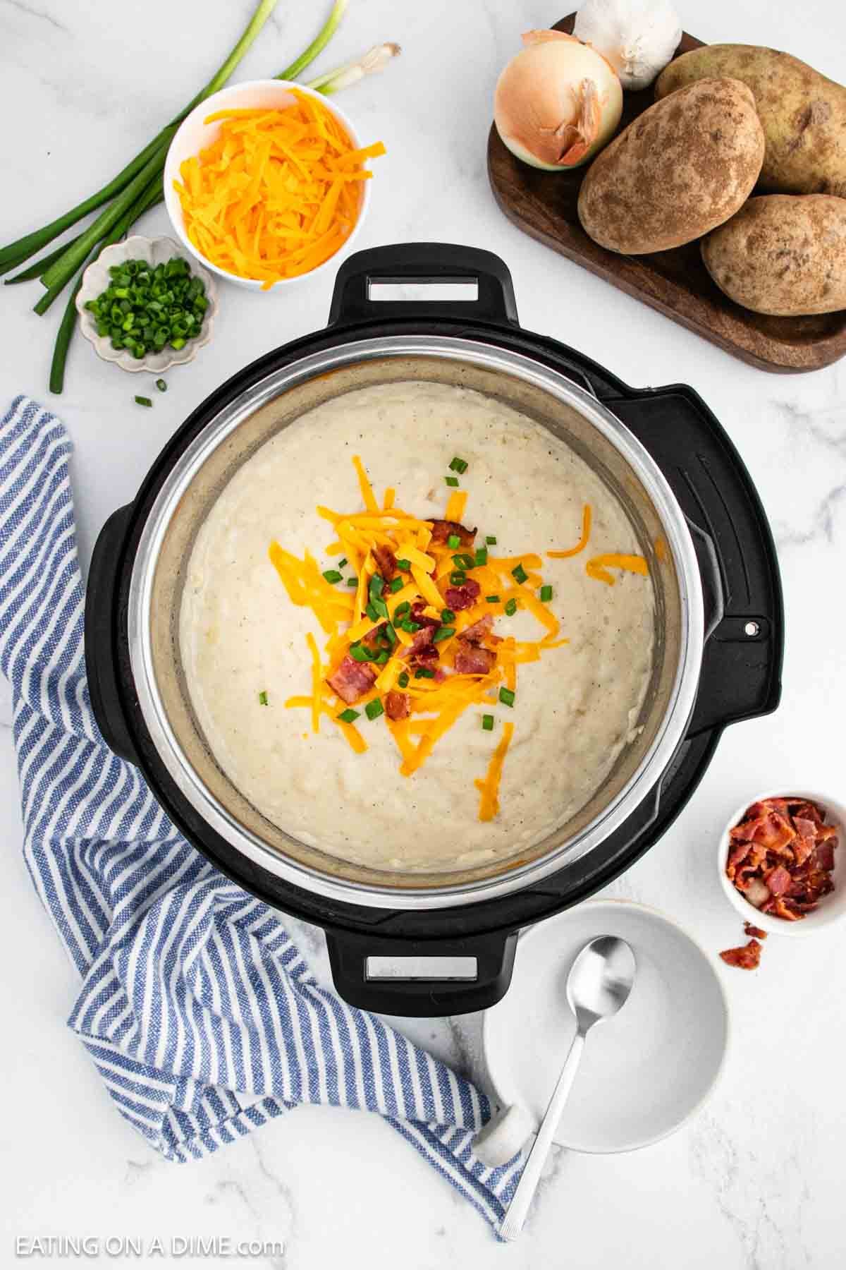 An overhead view of Instant Pot loaded baked potato soup, topped with shredded cheddar cheese, green onions, and bacon bits. Surrounding the pot are potatoes, cheese, green onions, bacon, and a blue striped towel.