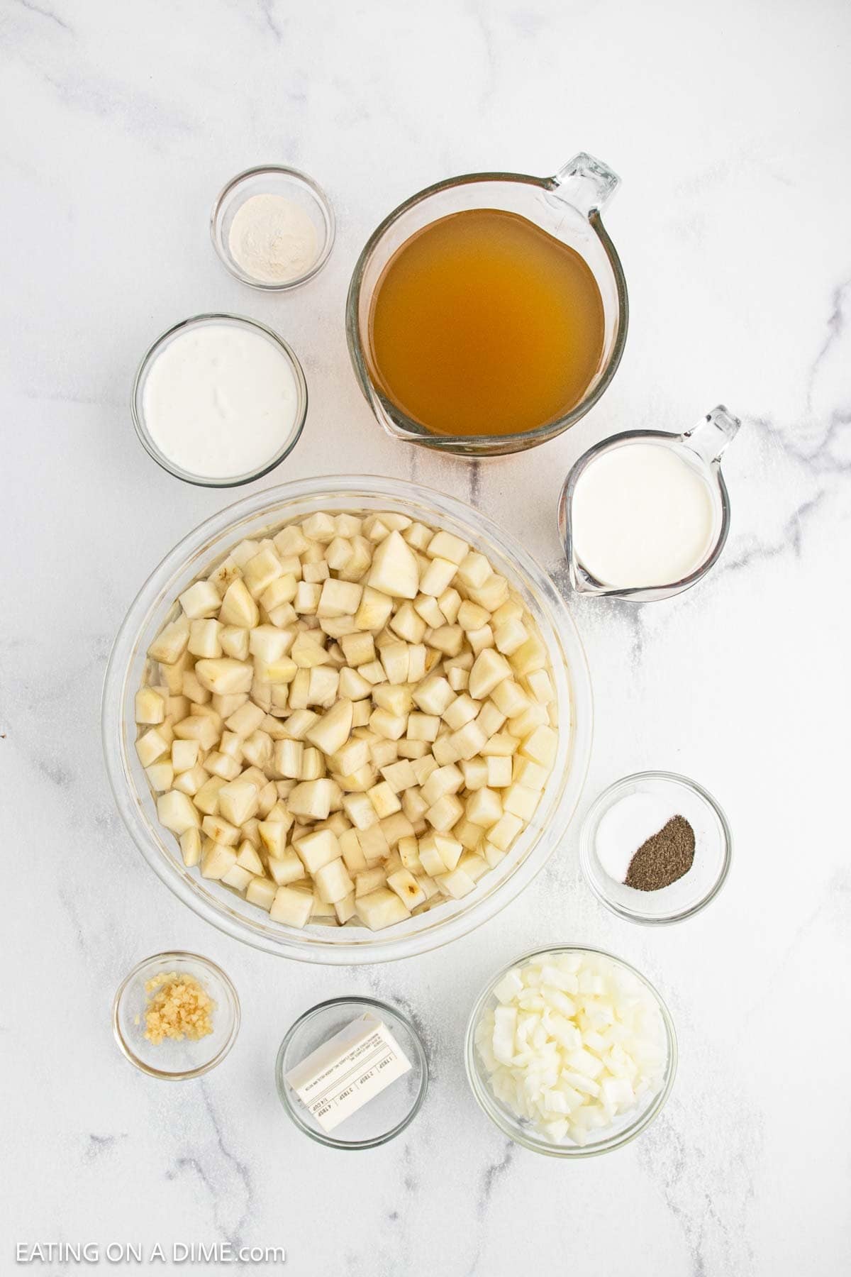 Overhead view of ingredients for Instant Pot loaded baked potato soup, featuring a large bowl of diced potatoes, broth in a measuring cup, cream, sour cream, diced onions, minced garlic, butter, pepper, and flour on a white surface.