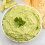 A bowl of creamy avocado dip guacamole garnished with a sprig of cilantro, served with tortilla chips and lime wedges on the side.