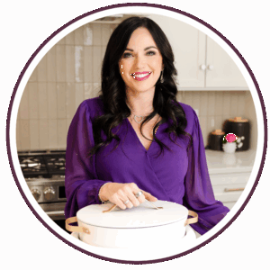 A woman with long dark hair, wearing a purple blouse, stands in a kitchen holding a white pot with a lid, smiling at the camera. The kitchen background includes a stove and decor items on the counter.
