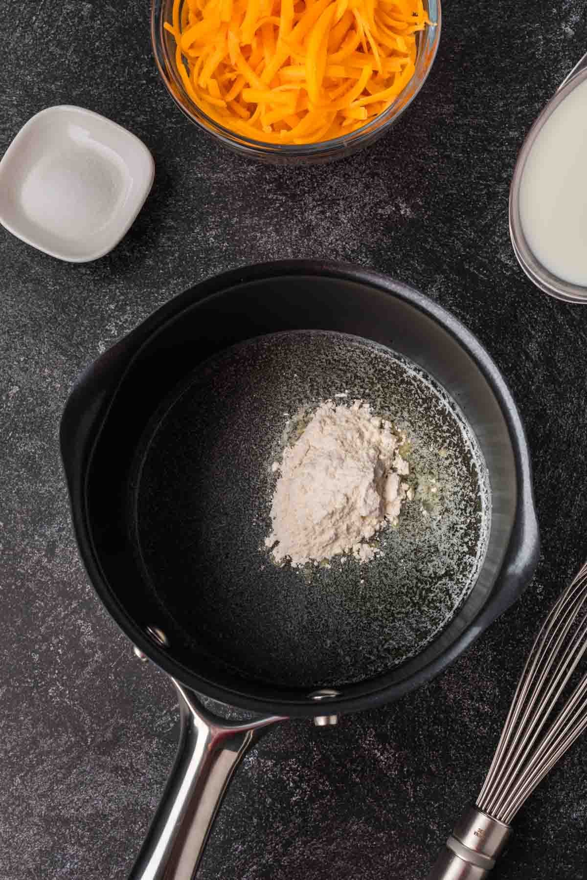 A black saucepan with melted butter and flour sits on a dark countertop, ready to become a rich Cheese Sauce for Pretzels. Nearby are a bowl of shredded cheddar, a dish of salt, milk, and a metal whisk.