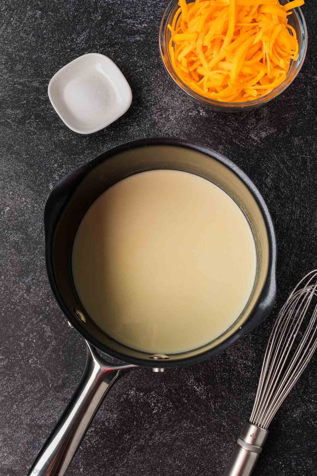 A saucepan with creamy white Cheese Sauce for Pretzels sits on a dark countertop next to a whisk, a small dish of salt, and a bowl of shredded cheddar cheese.