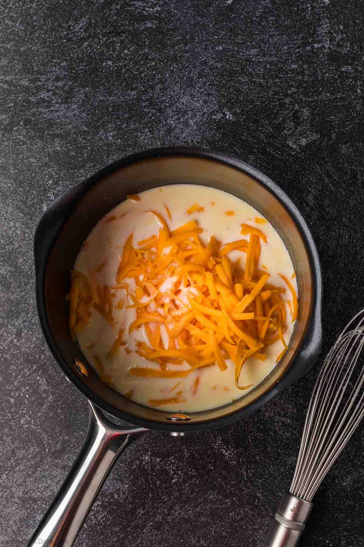 A saucepan filled with creamy Cheese Sauce for Pretzels and shredded cheddar cheese on top sits on a dark textured surface next to a metal whisk.