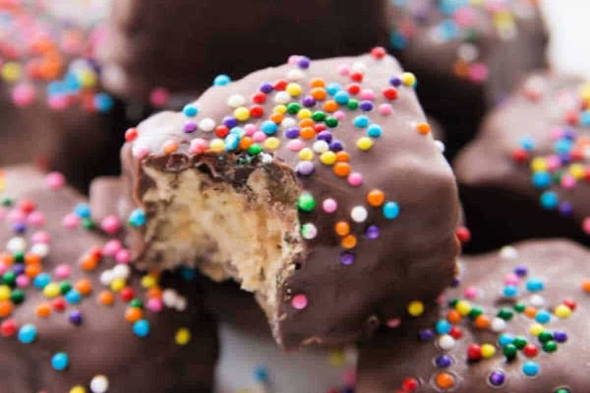 Try this delicious Chocolate Covered Rice Krispie Treats recipe. These chocolate dipped rice krispie treats are perfect for any Christmas cookie exchange, baby shower, wedding or birthday party. These DIY chocolate covered Rice Krispies are a quick and easy dessert idea. #eatingonadime #dessertrecipes A close-up of Chocolate Covered Rice Krispie Treats topped with colorful sprinkles, with one piece showing a bite revealing a creamy, light interior.