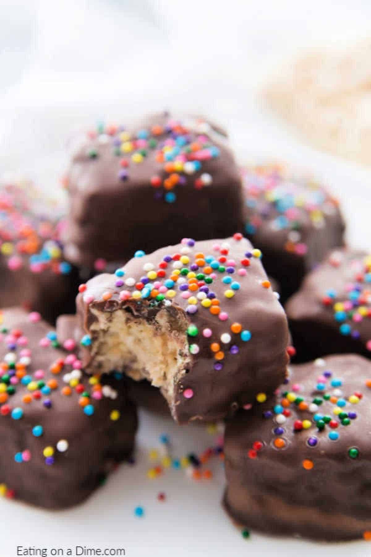 Try this delicious Chocolate Covered Rice Krispie Treats recipe. These chocolate dipped rice krispie treats are perfect for any Christmas cookie exchange, baby shower, wedding or birthday party. These DIY chocolate covered Rice Krispies are a quick and easy dessert idea. #eatingonadime #dessertrecipes A close-up of chocolate-covered rice krispie treats topped with colorful rainbow sprinkles. One treat has a bite taken out, revealing a creamy filling. They are arranged on a white surface.