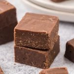 Two thick squares of 2 Ingredient Fudge are stacked on top of each other on a light gray surface, with more pieces of fudge and white plates visible in the background.