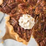 A slice of chocolate pecan pie topped with a dollop of whipped cream and chopped pecans, held above the rest of the pie with a pie server.