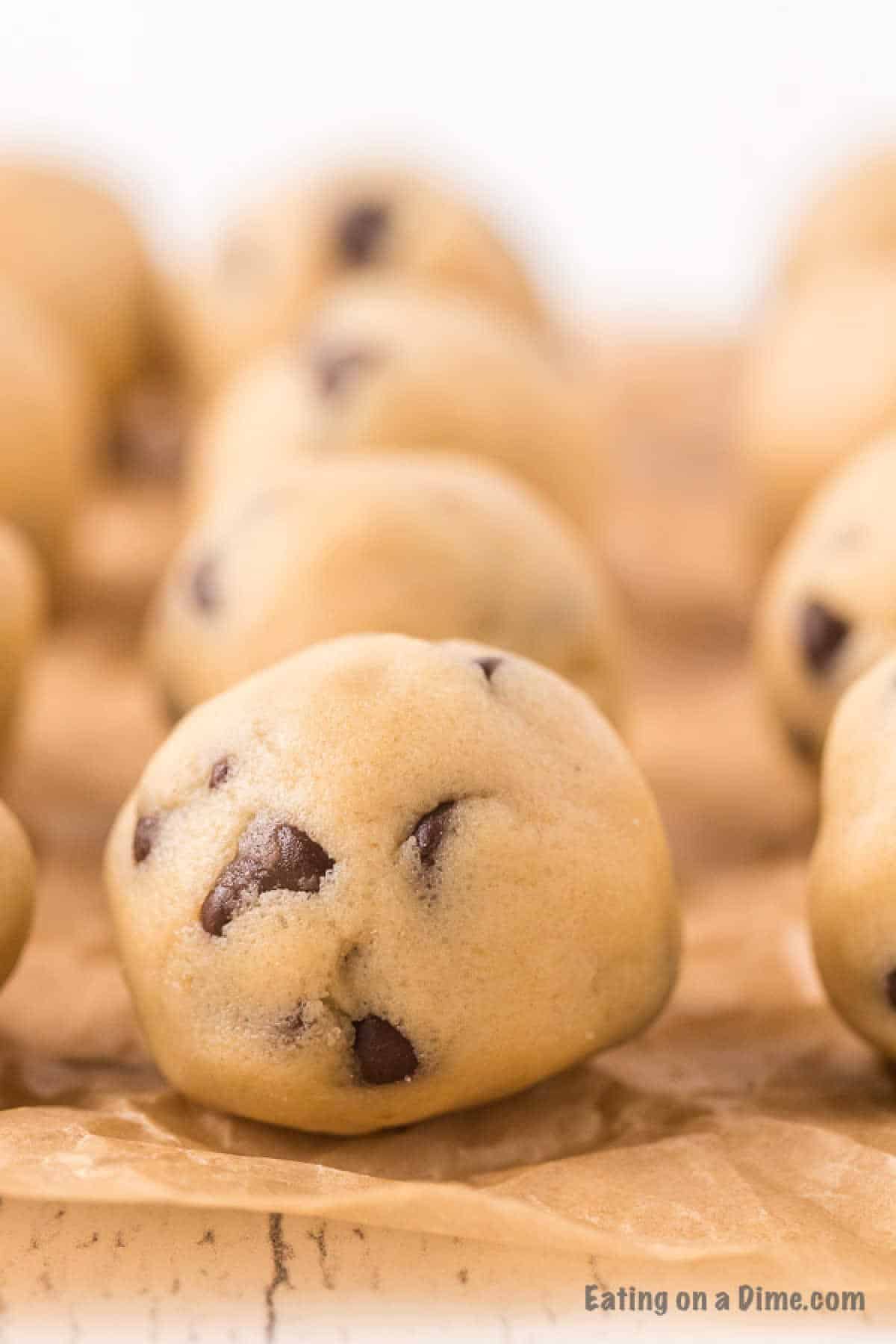 Edible cookie dough bites are arranged on parchment paper, ready for baking. The dough is a light brown hue with chocolate chips generously scattered throughout. A softly blurred background draws attention to the tempting bites in the foreground.