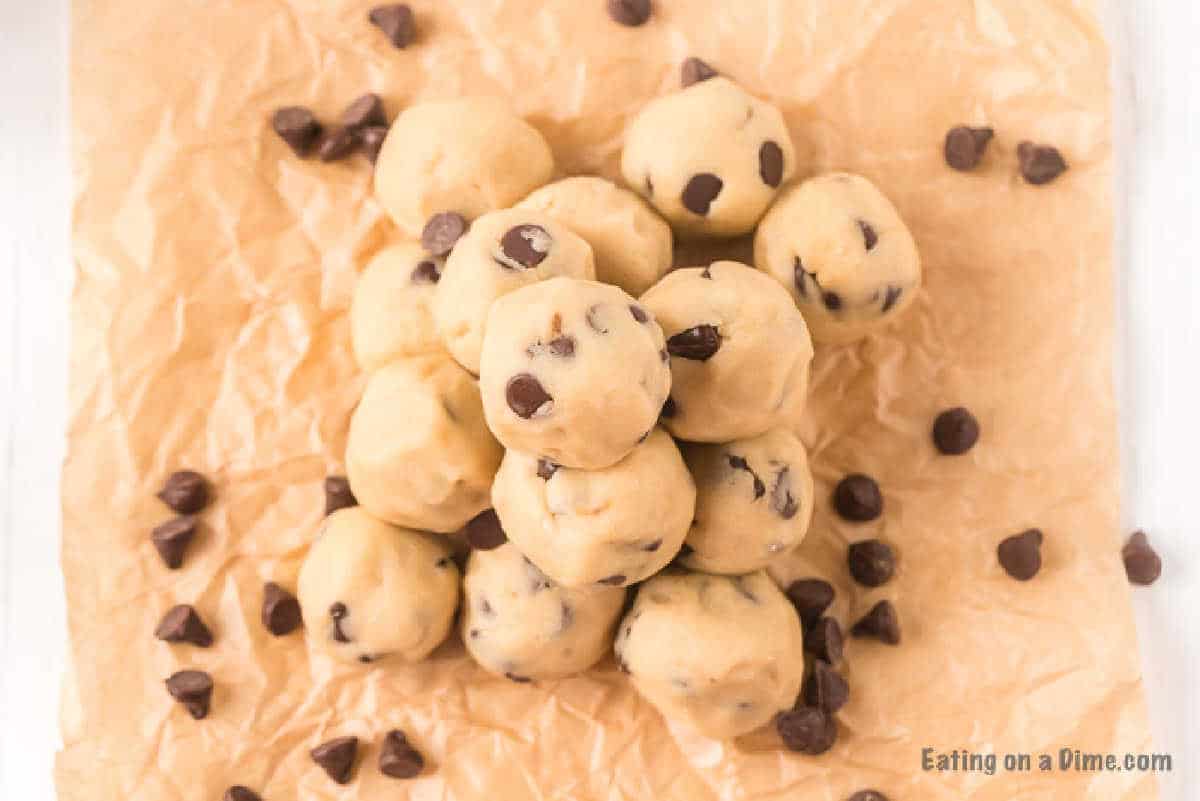 A pile of edible cookie dough bites is placed on crinkled parchment paper. A few scattered chocolate chips surround the dough balls, adding to the textured backdrop.