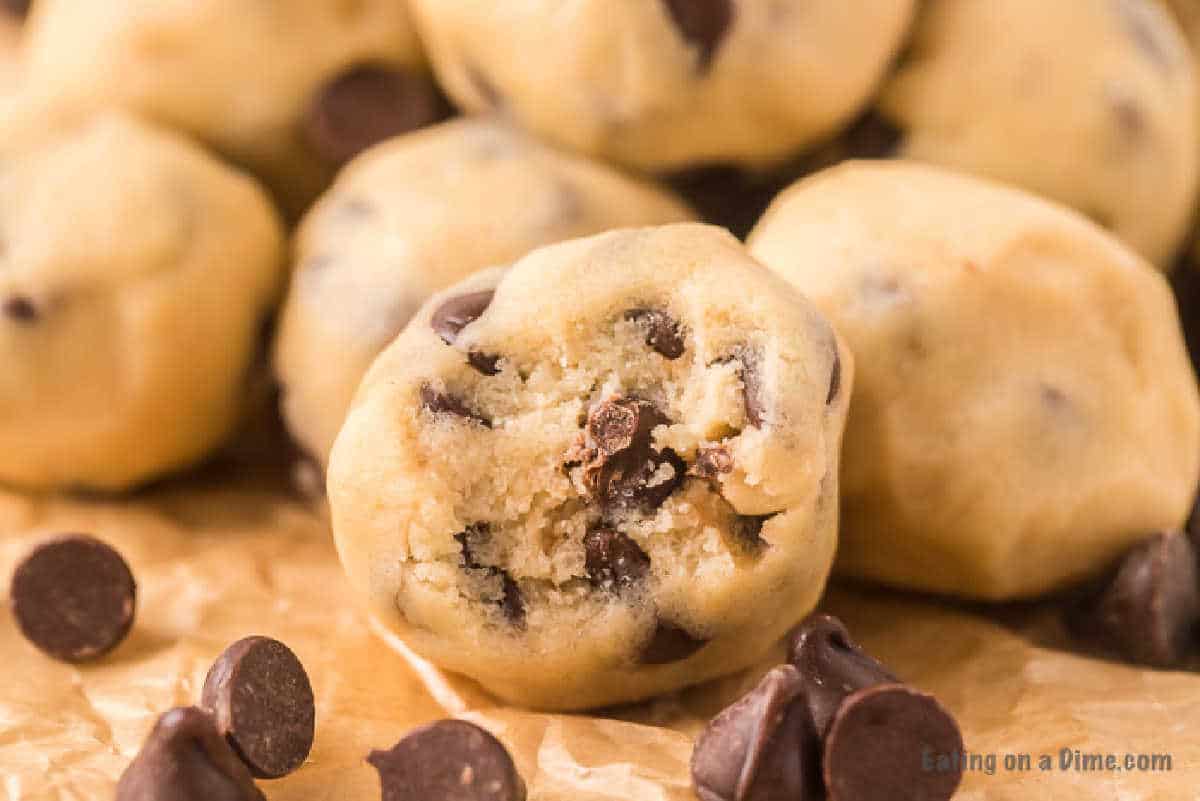 Close-up of edible cookie dough bites on parchment paper. One bite is prominently featured with chocolate chips visible inside. Other dough balls and scattered chocolate chips are in the background, highlighting their irresistible allure.