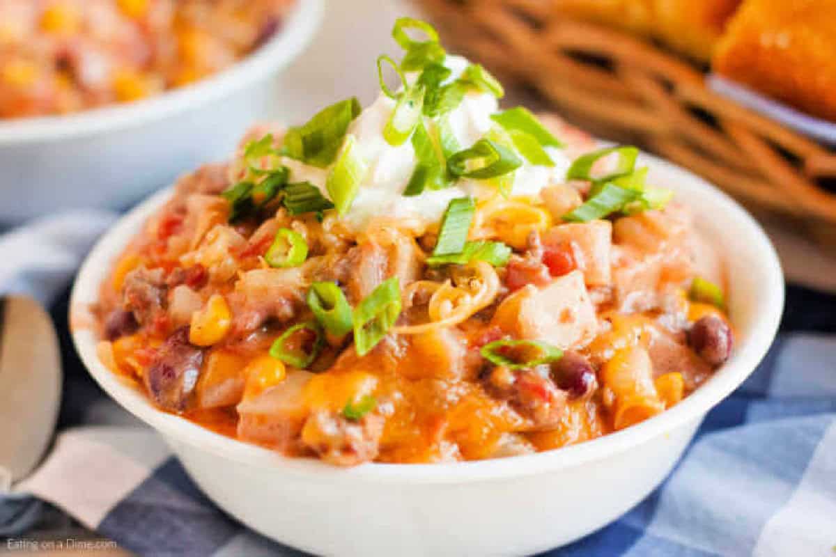 A bowl of creamy Crockpot Cowboy Casserole soup filled with beans, corn, tomatoes, and diced potatoes, topped with sour cream, shredded cheese, and chopped green onions. A basket of bread is blurred in the background.