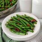 A plate of Green Beans and Bacon is served on a white dish, paired with a green checkered napkin and a fork. In the background, a skillet brimming with green beans and two white containers complete the inviting scene.