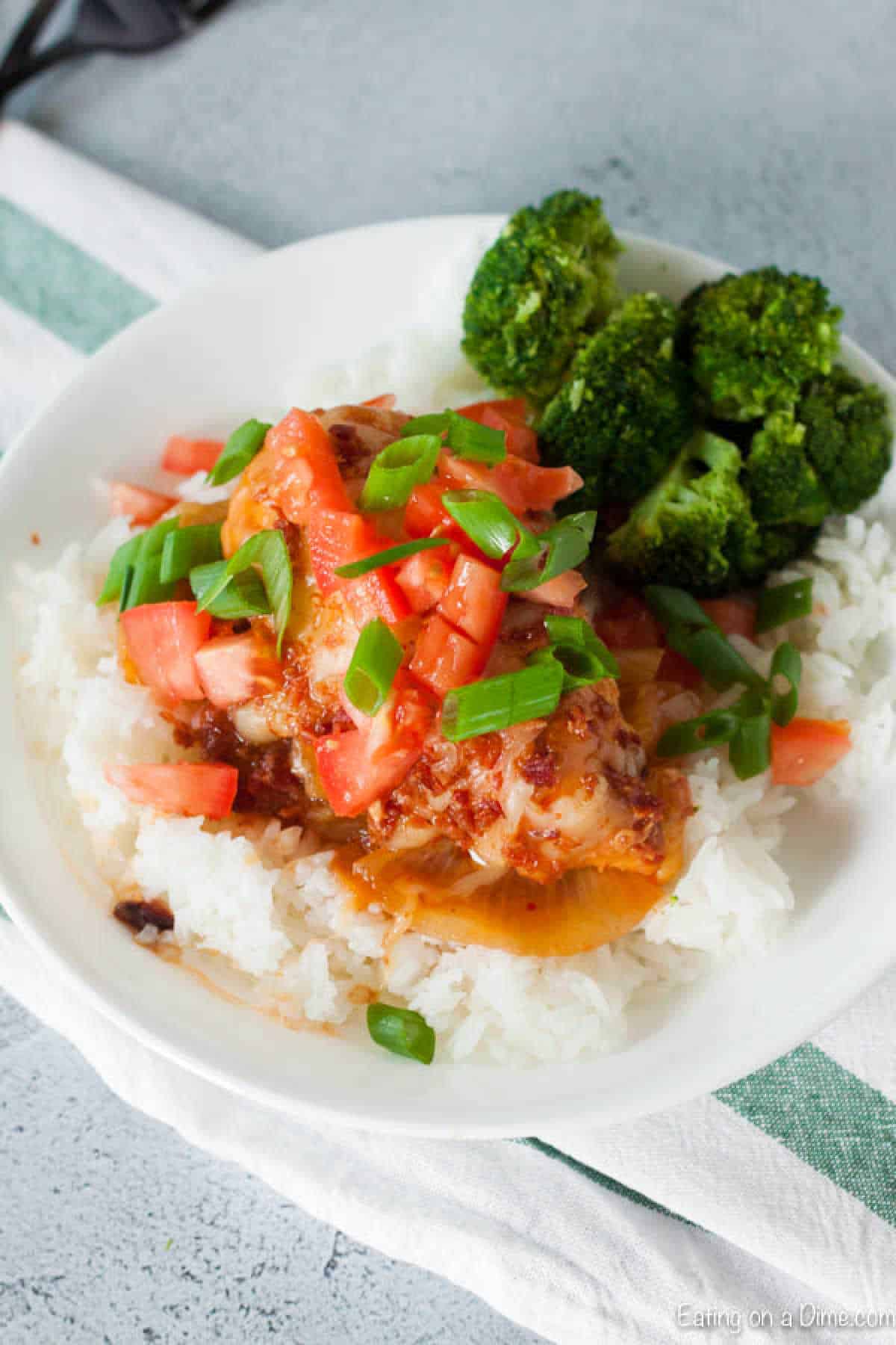 Try this crock pot monterey chicken recipe that is a copy cat Chili's recipe but even easier thanks to the slow cooker!ย Packed with BBQ sauce and cheese. A plate of rice topped with crock pot Monterey chicken, diced tomatoes, green onions, and sauce. The dish is accompanied by a side of steamed broccoli. A striped green and white napkin is partially visible underneath the plate.