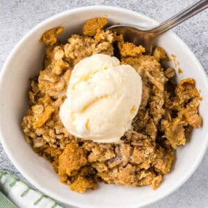 A white bowl filled with Pumpkin Crisp Recipe topped with vanilla ice cream, with a spoon resting in the bowl.