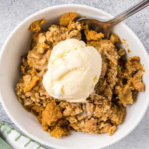 A white bowl filled with Pumpkin Crisp Recipe topped with vanilla ice cream, with a spoon resting in the bowl. The dessert features a crumbly oat topping and sits on a light surface next to a green napkin.