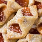 Close-up of several Strawberry Pinch Cookies, featuring golden, flaky pastry filled with red fruit jam and dusted with powdered sugar.