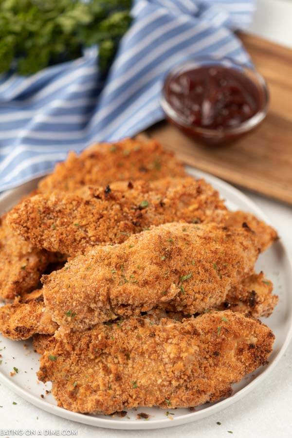 Lots of baked breaded chicken strips on a white plate topped with fresh parsley with a small bowl of BBQ sauce behind it.  