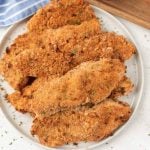 A plate stacked with golden-brown, oven baked breaded chicken tenders garnished with herbs. The tenders look crispy and freshly baked, with crumbs scattered around the plate.