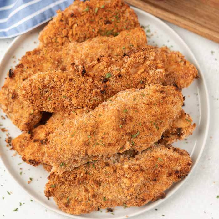 Lots of baked breaded chicken strips on a white plate topped with fresh parsley on top of them 