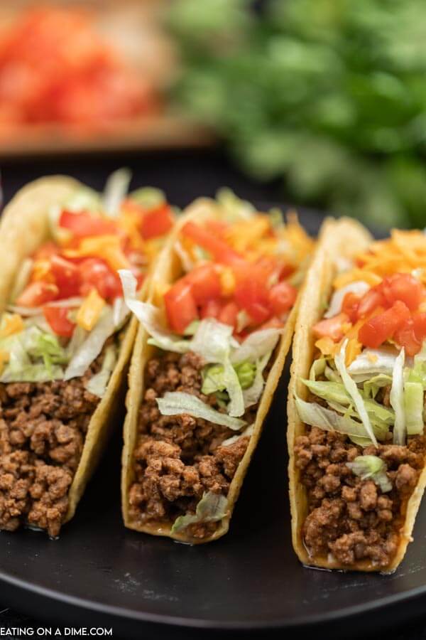 close up image of three tacos on a black plate. they are topped with lettuce, cheese and tomatoes. 