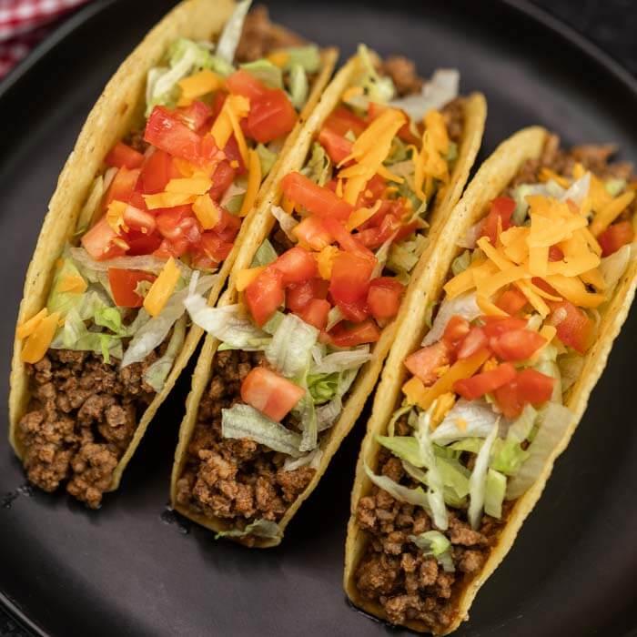 Close up image of three tacos on a black plate. The tacos are topped with lettuce, cheese and tomatoes. 