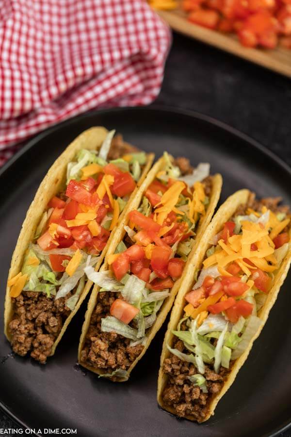 Close up image of three tacos on a black plate. They are topped with lettuce, cheese and tomatoes. 