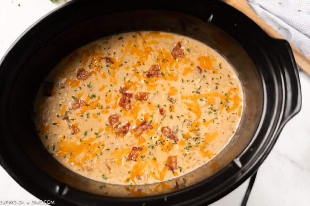 A creamy Crockpot Crack Potato Soup with shredded cheddar cheese, bacon bits, and herbs sprinkled on top, served in a black slow cooker.