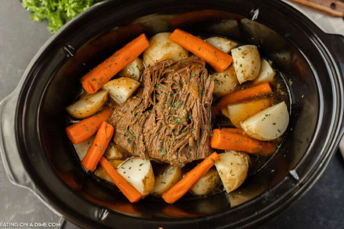 A Crock Pot Roast simmers in a slow cooker, surrounded by sliced carrots and potato wedges, all beautifully garnished with fresh herbs.
