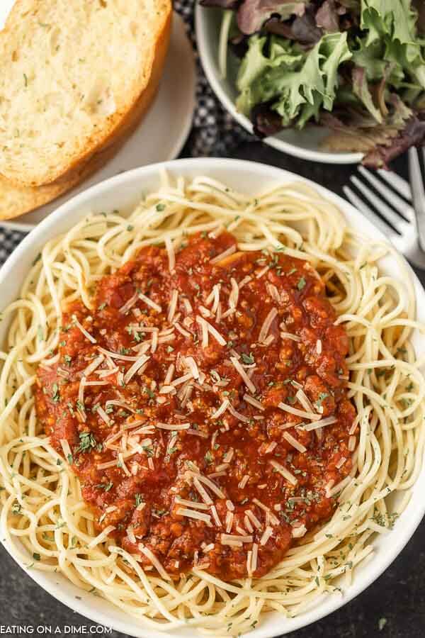 Close up image of a spaghetti with sauce on top on a white plate with a side of french bread and salad. 