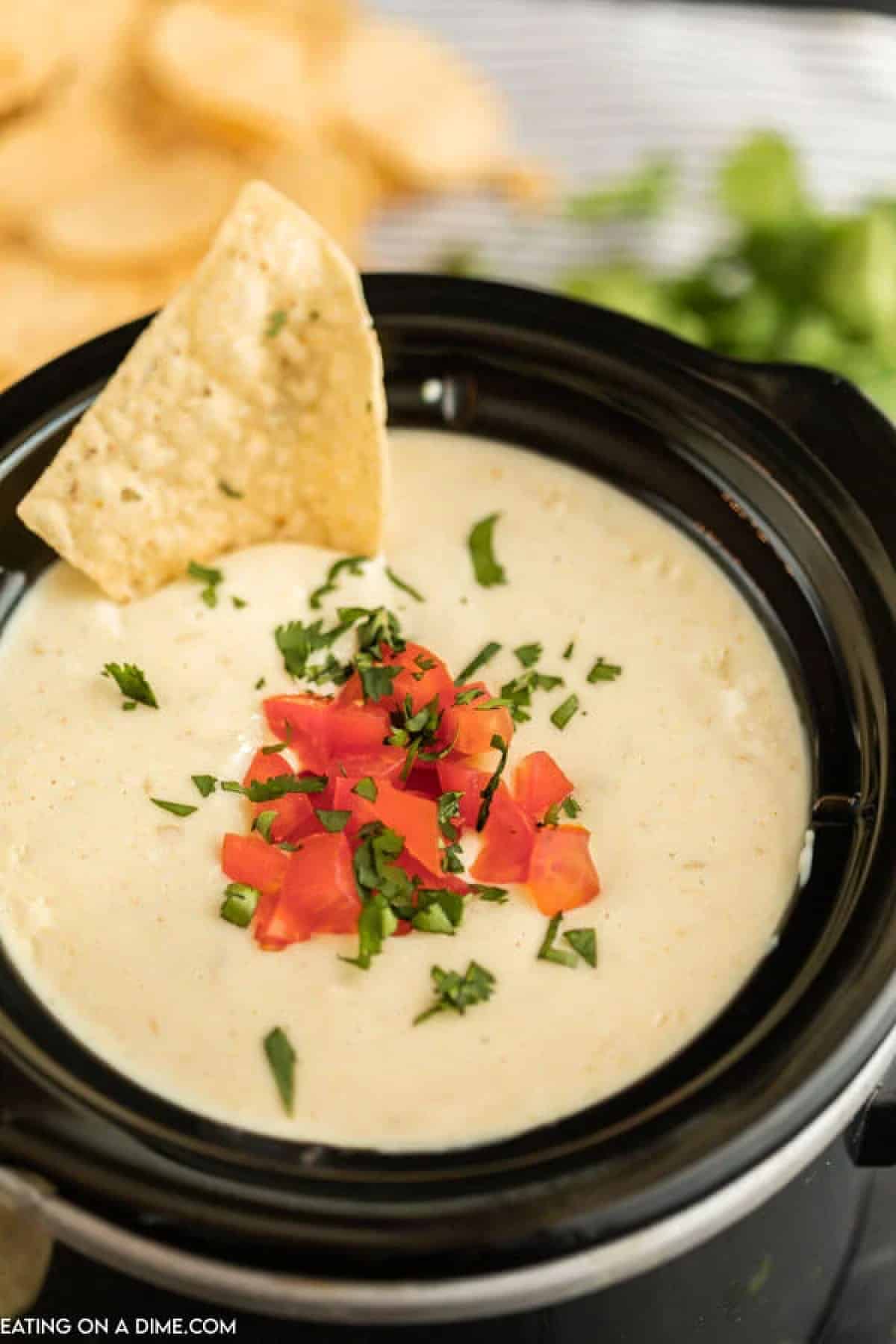 White Cheese Dip in a small crock pot topped with diced tomatoes, cilantro and a chip dipped into it.  
