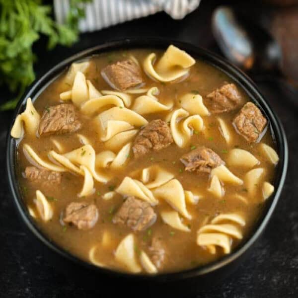 A black bowl filled with slow cooker steak soup, featuring chunks of beef and wide egg noodles in a brown broth, sits on a dark surface with fresh herbs in the background.