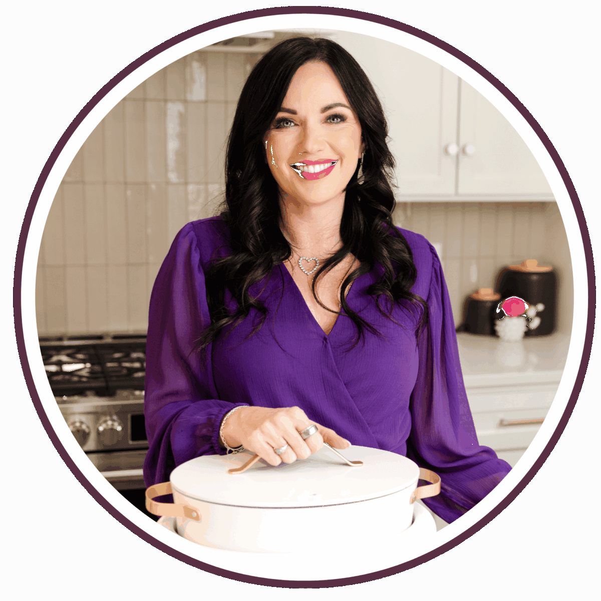 A woman with long dark hair wearing a purple blouse stands in a kitchen, smiling at the camera while holding a white pot with a lid. The background shows a stove, cabinets, and decorative items.