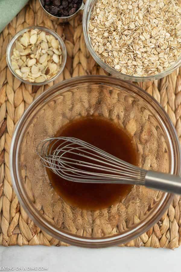 Close up image of the oil and honey being mixed in a bowl with the oats, sliced almonds and raisins on the side. 