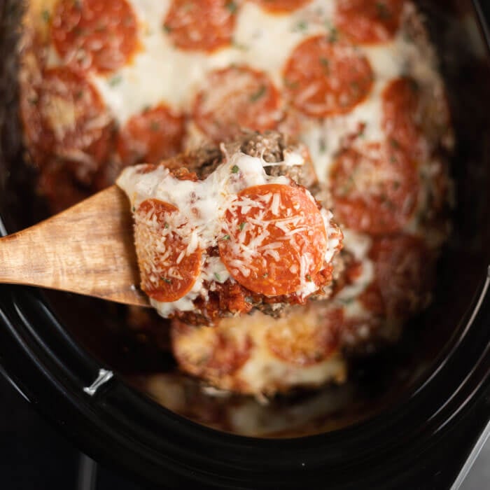Close up image of pizza casserole in a crock pot with a serving on a wooden spoon. 