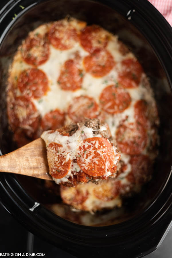 Close up image of pizza casserole in the crock pot with a serving on a wooden spoon. 