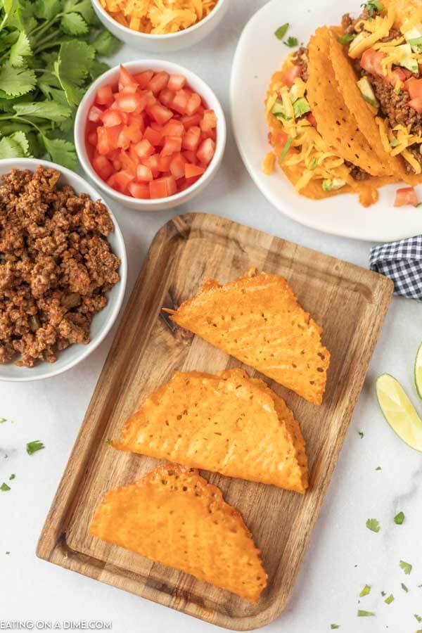 Close up image of keto taco shells on a cutting board with a side of hamburger meat, tomatoes and cheese. With a white plate of tacos. 