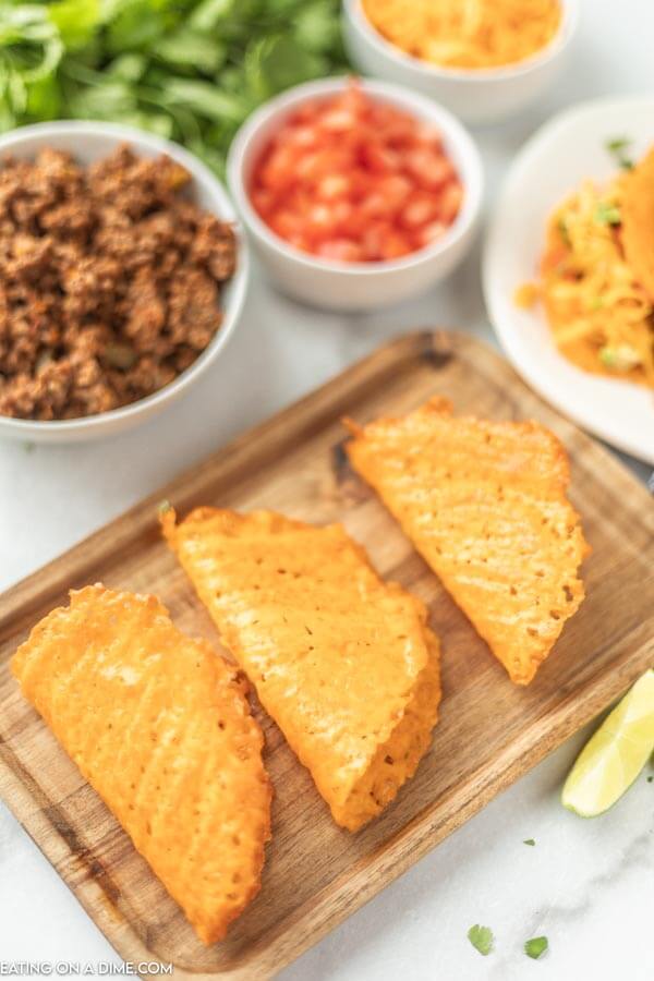 Close up image of keto taco shells on a cutting board with a side of hamburger meat, tomatoes, and cheese. 