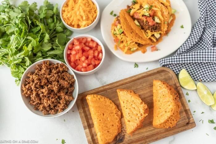 Close up image of keto taco shells on a platter, with a side of hamburger meat, tomatoes and cheese. Also a white plate with tacos. 