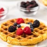 A golden Keto Waffles Recipe topped with raspberries, blackberries, blueberries, and a pat of butter, with syrup drizzled on the side. Blurred bowls of berries are in the background.