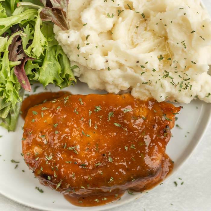 Close up image of pork chops covered in marinade with a side of mashed potatoes and salad on a white plate. 